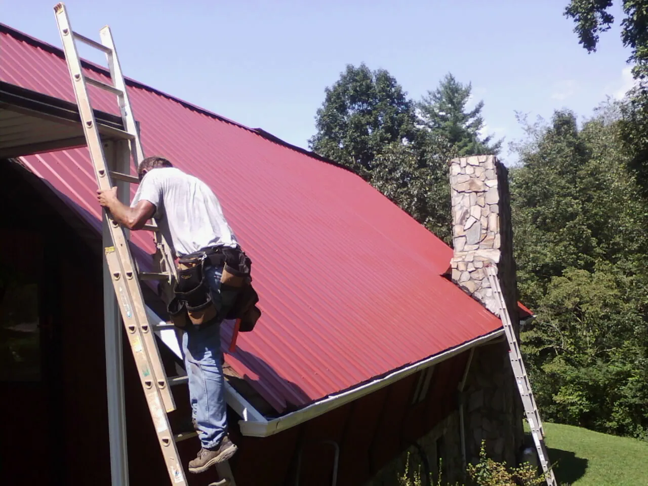 Roofer on a ladder beside a red metal roof and stone chimney — Soffit & Fascia Repair by Roofing Brothers in Martinsville